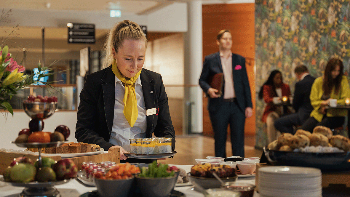 Femme souriante préparant un buffet de pause dans un espace de conférence. Homme en costume marchant en arrière-plan.
